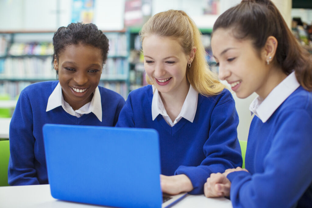 Three smiling female students wearing blue school uniforms working on laptop in library